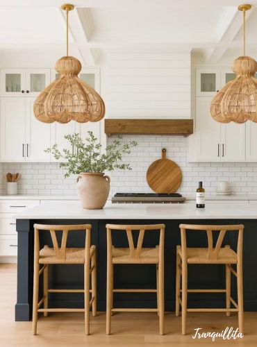 A bright, coastal-style kitchen white cabinets blend in with subway tile white walls to create seamless look and a rustic wood vent hood create a clean, airy look. Light wood wishbone-style bar stools under dark navy blue island with a white countertop and two large pendant lights overhead. Transformed kitchen by Tranquillita of a cramped 2 BHK near Alipur Mint