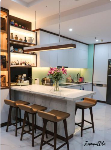 A modern kitchen with a white dining bar, a long, minimalist light fixture hangs from the ceiling above it. Shelving unit with built-in lighting to the left. A tidy kitchen counter. 2 BHK at Sonarpur Mission Pally by Tranquillita.